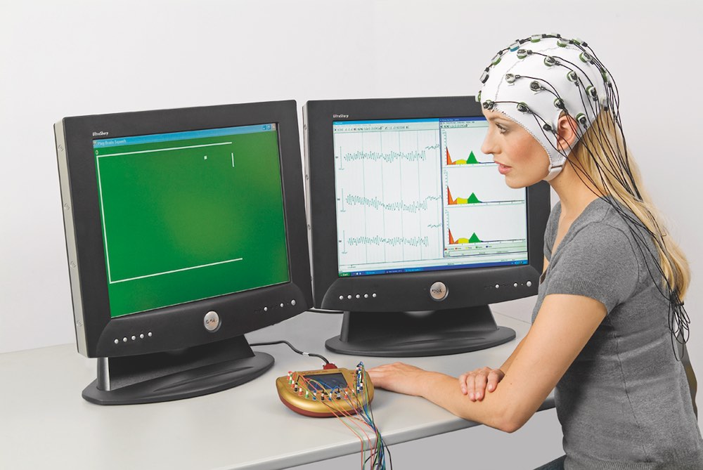 An EEG system. The model is wearing a cap to which multiple electrodes are attached. The electrodes connect to the gold-colored amplifier sitting on the table. The right screen shows the ongoing EEG recorded from the scalp, while the left screen shows the experimental stimuli that the model is attending to. Image courtesy Brain Vision LLC.